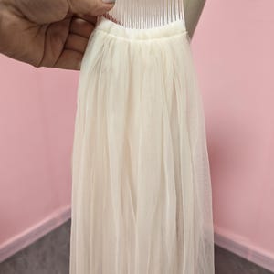 May include: A sheer, off-white tulle veil attached to a clear plastic hair comb. The fabric is gathered at the top, creating soft, vertical folds that drape downward. The lightweight material is translucent and features a simple, unadorned design.