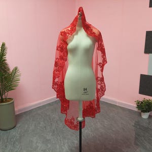 May include: A red bridal veil with floral lace detailing is draped over a mannequin. The veil is sheer, allowing the mannequin to be seen through it. The lace is concentrated along the edges and around the top of the veil.