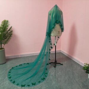 May include: A long, floor-length emerald green veil made of sheer tulle fabric. The veil is draped over a dress form and features numerous small, dark green butterfly-shaped appliqu&eacute;s attached along the edges and scattered across the fabric.