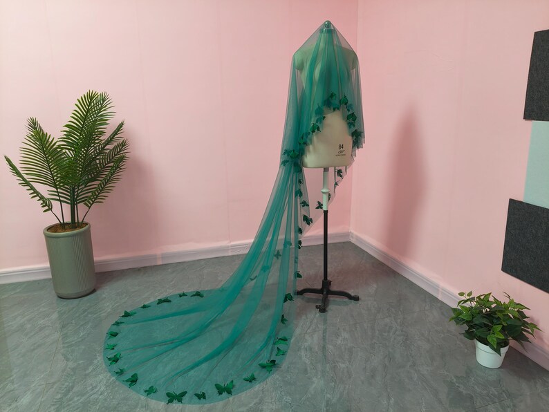 May include: A long, floor-length emerald green veil made of sheer tulle fabric. The veil is draped over a dress form and features numerous small, dark green butterfly-shaped appliqu&eacute;s attached along the edges and scattered across the fabric.
