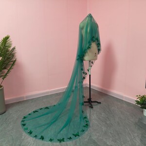 May include: A long, floor-length emerald green veil made of sheer tulle fabric. The veil is draped over a mannequin and features numerous small, dark green butterfly-shaped appliqu&eacute;s scattered along the edges and throughout the length of the fabric.