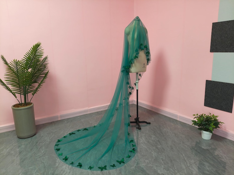 May include: A long, floor-length emerald green veil made of sheer tulle fabric. The veil is draped over a mannequin and features numerous small, dark green butterfly-shaped appliqu&eacute;s scattered along the edges and throughout the length of the fabric.