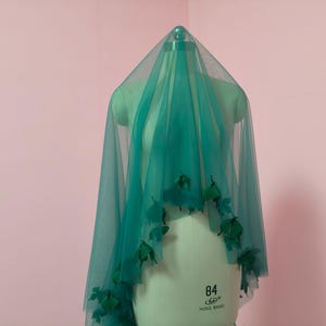 May include: A sheer teal tulle veil draped over a mannequin, featuring dark green fabric leaf embellishments attached along the lower edges. The veil is translucent, creating a layered effect as it hangs from the top of the mannequin form.
