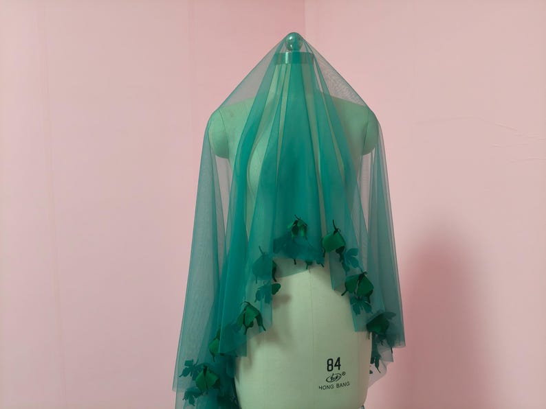 May include: A sheer teal tulle veil draped over a mannequin, featuring dark green fabric leaf embellishments attached along the lower edges. The veil is translucent, creating a layered effect as it hangs from the top of the mannequin form.