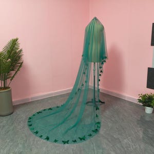 May include: A long, floor-length teal green veil made of sheer tulle fabric. The veil is draped over a mannequin and features numerous small, three-dimensional butterfly embellishments attached along the edges and scattered across the fabric.