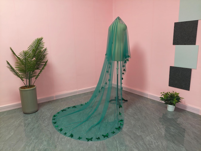 May include: A long, floor-length teal green veil made of sheer tulle fabric. The veil is draped over a mannequin and features numerous small, three-dimensional butterfly embellishments attached along the edges and scattered across the fabric.