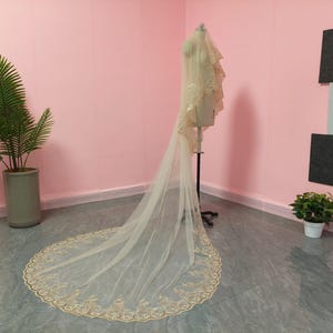 Può includere: Un lungo velo da sposa avorio con un bordo ricamato dorato dettagliato. Il velo è esposto su un manichino, mostrando la sua lunghezza e il suo design delicato. Lo strascico del velo si estende sul pavimento.