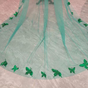 May include: A long, sheer, emerald green tulle veil featuring a rounded hemline. The edges of the veil are decorated with numerous three-dimensional, solid emerald green butterfly appliques of varying sizes, arranged along the curved perimeter.