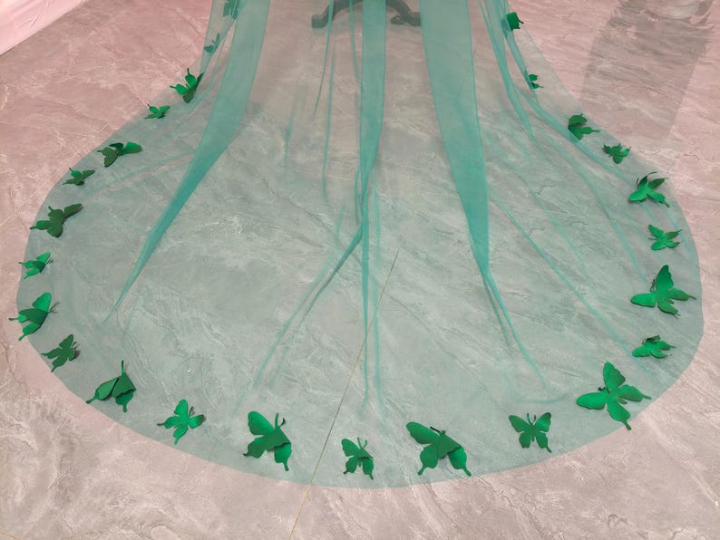 May include: A long, sheer, emerald green tulle veil featuring a rounded hemline. The edges of the veil are decorated with numerous three-dimensional, solid emerald green butterfly appliques of varying sizes, arranged along the curved perimeter.
