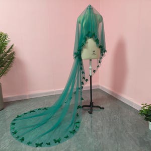 May include: A long, floor-length emerald green veil made of sheer tulle fabric. The veil is draped over a dress form and is decorated with numerous small, dark green butterfly-shaped appliqu&eacute;s scattered along the edges and throughout the length.