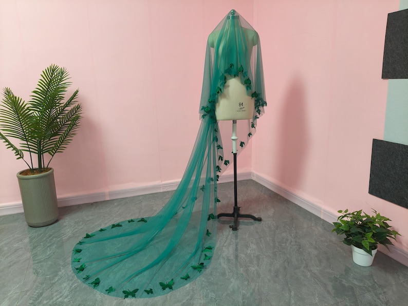 May include: A long, floor-length emerald green veil made of sheer tulle fabric. The veil is draped over a dress form and is decorated with numerous small, dark green butterfly-shaped appliqu&eacute;s scattered along the edges and throughout the length.