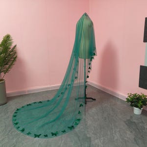 May include: A long, floor-length emerald green veil made of sheer tulle fabric. The veil is draped over a mannequin and features numerous small, dark green butterfly-shaped appliqu&eacute;s attached along the edges and scattered throughout the fabric.