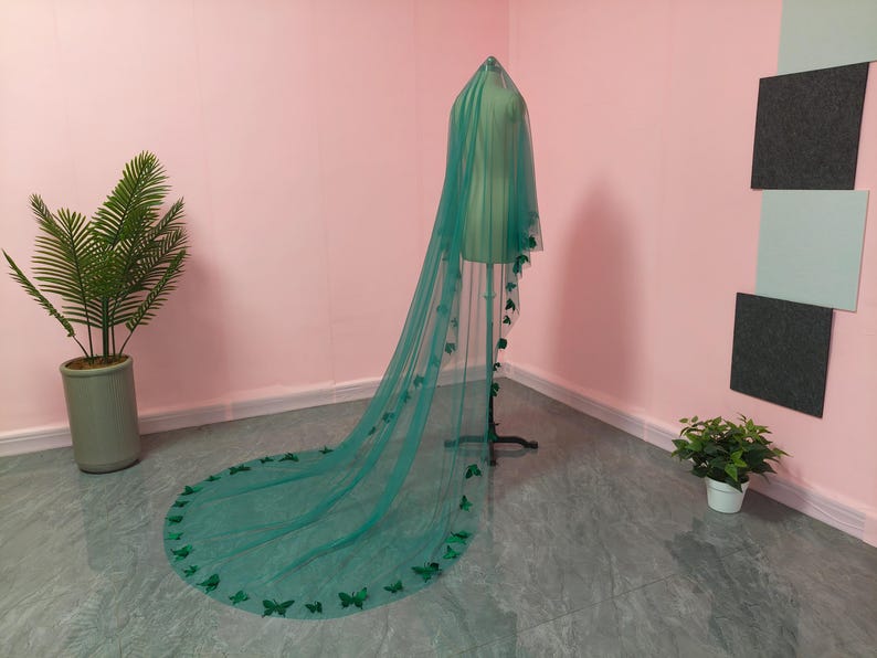 May include: A long, floor-length emerald green veil made of sheer tulle fabric. The veil is draped over a mannequin and features numerous small, dark green butterfly-shaped appliqu&eacute;s attached along the edges and scattered throughout the fabric.