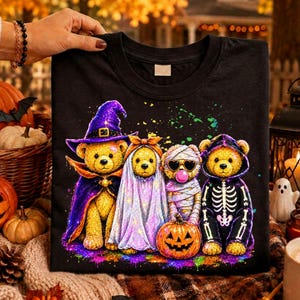 以下が含まれることがあります： ハロウィンの仮装をした4匹のテディベアが描かれた黒いTシャツ。1匹は紫の魔女の帽子とマントを身につけ、1匹は幽霊、1匹はハート型のサングラスをかけたミイラ、もう1匹はカボチャの横で骸骨の衣装を着ている様子がプリントされています。