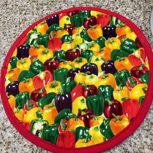May include: A round fabric pot holder featuring a vibrant, all-over print of colorful bell peppers in shades of red, yellow, green, orange, and purple. The circular mat is finished with a solid red fabric binding along the outer edge.