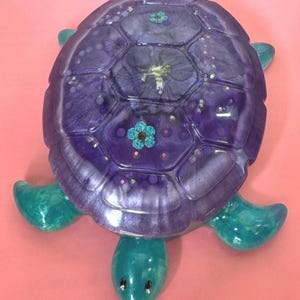 May include: A turtle-shaped decorative object made of translucent resin. The shell is a deep purple with embedded blue dried flowers and iridescent glitter. The head and four flippers are a shimmering teal colour with a smooth, glossy finish.