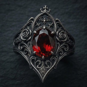 May include: A dark silver-toned ring featuring an oval-cut deep red gemstone at the center. The metal band is intricately detailed with sculpted rose motifs, scrolling filigree patterns, and a small bat silhouette positioned above the stone.