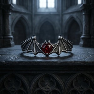 May include: A silver-toned ring in the shape of a bat with outstretched wings. The ring features a large, round, red gemstone at the center. The wings have detailed veining. The ring is set against a dark, gothic-style background.