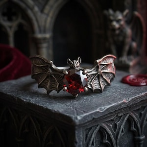May include: A silver-toned ring featuring a bat design with outstretched wings. The ring has a large, faceted, red gemstone at its center. The wings are detailed with vein patterns, and the bat's head is at the top of the gemstone.