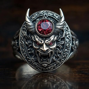 May include: A silver-colored ring featuring a detailed demonic face design with horns and a central red gemstone. The ring has intricate carvings and a dark, antique finish, showcasing a gothic or fantasy theme.