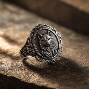 May include: A silver-toned ring featuring an oval centrepiece with a detailed relief of a wolf head. The band and the frame surrounding the wolf are decorated with intricate, raised floral and scrollwork patterns, finished with a dark patina.