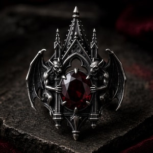 May include: A silver ring featuring a large, oval-cut dark red gemstone set within an intricate architectural frame. Two winged gargoyle figures are positioned on either side of the stone, integrated into the metalwork of the pointed arch structure.