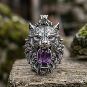May include: A detailed silver pendant depicting a wolf's head with an open mouth, showcasing a large, oval, purple gemstone. The wolf's fur is intricately carved, and the pendant has a loop for a chain.