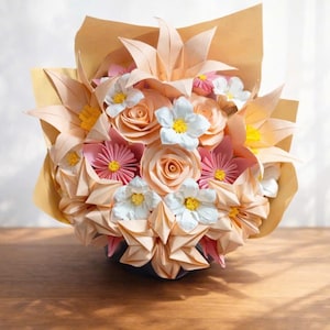 Könnte beinhalten: Ein Strauß Papierblumen in Pfirsich-, Rosa- und Weißtönen. Das Arrangement umfasst Rosen, Lilien und Gänseblümchen, alle aus gefaltetem Papier gefertigt. Der Strauß ist in braunes Papier gewickelt und schafft eine einzigartige und künstlerische Blumendarstellung.