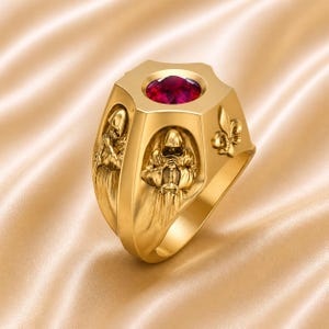 Könnte beinhalten: Ein goldfarbener Ring mit einem großen, facettierten, roten Edelstein in einer sechseckigen Lünette. Der Ring zeigt detaillierte Reliefs von Figuren in Kapuzenroben und ein Fleur-de-Lis-Design. Der Ring hat eine polierte Oberfläche und ein breites Band.
