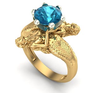May include: A gold-colored ring featuring two mermaid figures supporting a large, round, blue gemstone. The mermaids are intricately detailed, with scaled tails and flowing hair. The ring's band is smooth and polished.