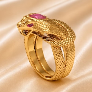 Könnte beinhalten: Ein goldfarbener Ring in Form einer Schlange. Der Schlangenkopf hat einen großen rosa Edelstein und kleinere rosa Edelsteine für die Augen. Der Ring hat ein strukturiertes, schuppenartiges Muster und ein Doppelband-Design.