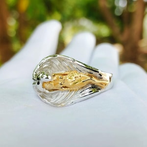 May include: A two-tone ring featuring a gold figure within a silver wave design. The ring is held in a white gloved hand, with a blurred green and brown background.
