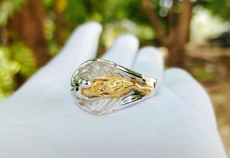 May include: A two-tone ring featuring a gold figure within a silver wave design. The ring is held in a white gloved hand, with a blurred green and brown background.
