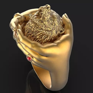 May include: A gold-toned ring featuring a sculpted design of two hands cupping a roaring lion head. The band includes a small, circular red gemstone accent embedded on the side, with detailed textures on the lion's mane and the fingers of the hands.