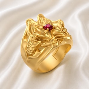 Könnte beinhalten: Ein goldfarbener Ring mit einem aufwendigen Design, das geformte Figuren und einen zentralen roten Edelstein aufweist. Der Ring hat ein breites Band und detaillierte Reliefarbeiten, die einen komplexen, verzierten Stil zeigen. Der Edelstein ist tiefrot.