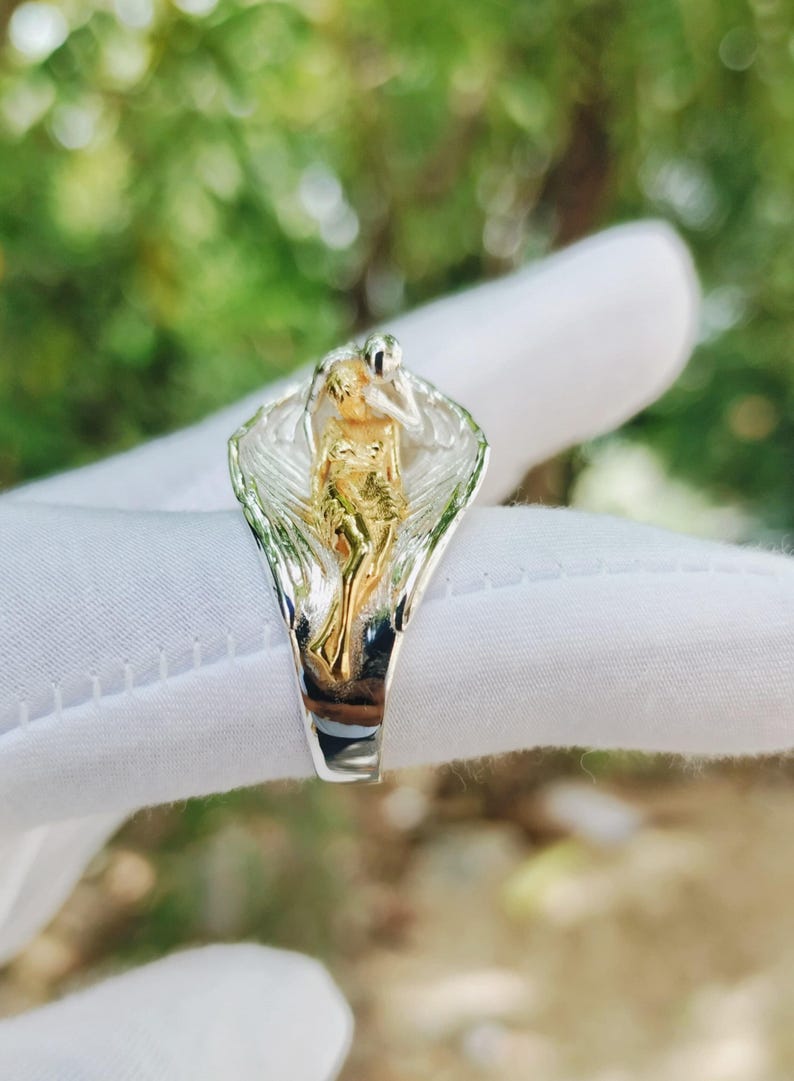 May include: A silver ring featuring a gold-toned figure within a stylized design. The ring's design includes a figure with outstretched arms, set against a backdrop of silver and gold. The ring is shown against a blurred green background.