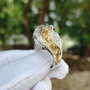 May include: A silver ring with a gold-colored figure of a person. The ring has a detailed design with a textured appearance. The background is blurred, with green and brown tones.
