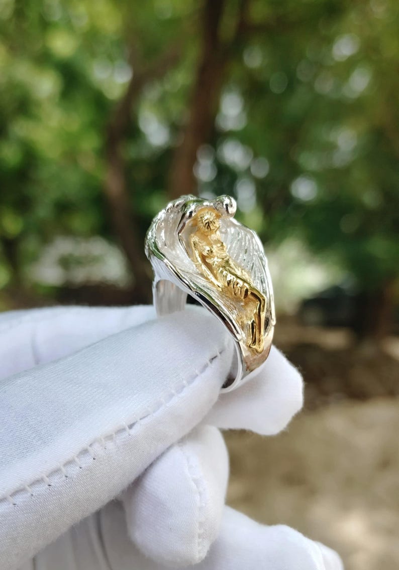 May include: A silver ring with a gold-colored figure of a person. The ring has a detailed design with a textured appearance. The background is blurred, with green and brown tones.