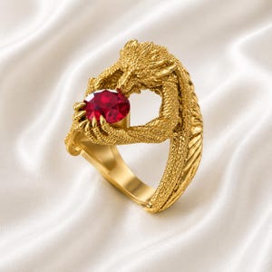 Peut inclure: Une bague dorée ornée d'un dragon tenant une grosse pierre rouge ovale. Les griffes et la tête du dragon entourent la pierre précieuse. Le fond blanc et soyeux met en valeur les détails de l'anneau, créant un bijou unique.