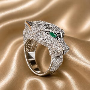 Könnte beinhalten: Silberfarbener Ring mit Pantherkopf-Design, besetzt mit funkelnden, klaren Steinen. Die Augen des Panthers sind smaragdgrün. Der Ring ist vor einem goldfarbenen, satinartigen Hintergrund platziert.