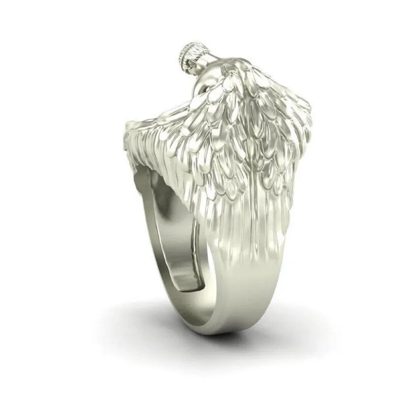 May include: A silver-colored ring featuring a detailed angel wing design. The ring has a sculpted angel figure at the top, with the wings extending down the sides. The ring is shown against a white background.