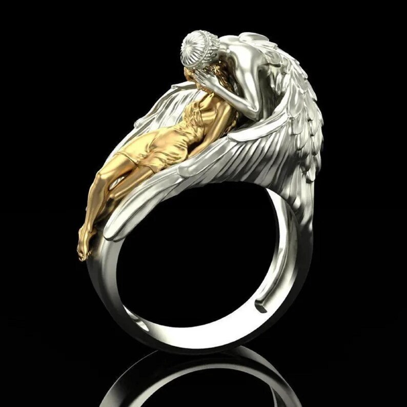 May include: A silver ring featuring a detailed sculpture of two figures. One figure is gold-colored and reclining, while the other, silver-colored, appears to be embracing the first. The ring has a polished finish and a wide band, set against a black background.