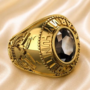 May include: A gold-colored ring with the words "UNITED STATES ARMY" encircling a dark, oval-shaped gemstone. The ring has intricate side detailing, including a crest and wings, set against a satin background.