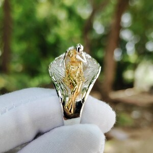May include: A two-tone ring featuring a gold-colored figure within silver-colored wings. The ring is held by a gloved hand against a blurred green background. The ring's design is intricate and detailed, showcasing a unique artistic style.