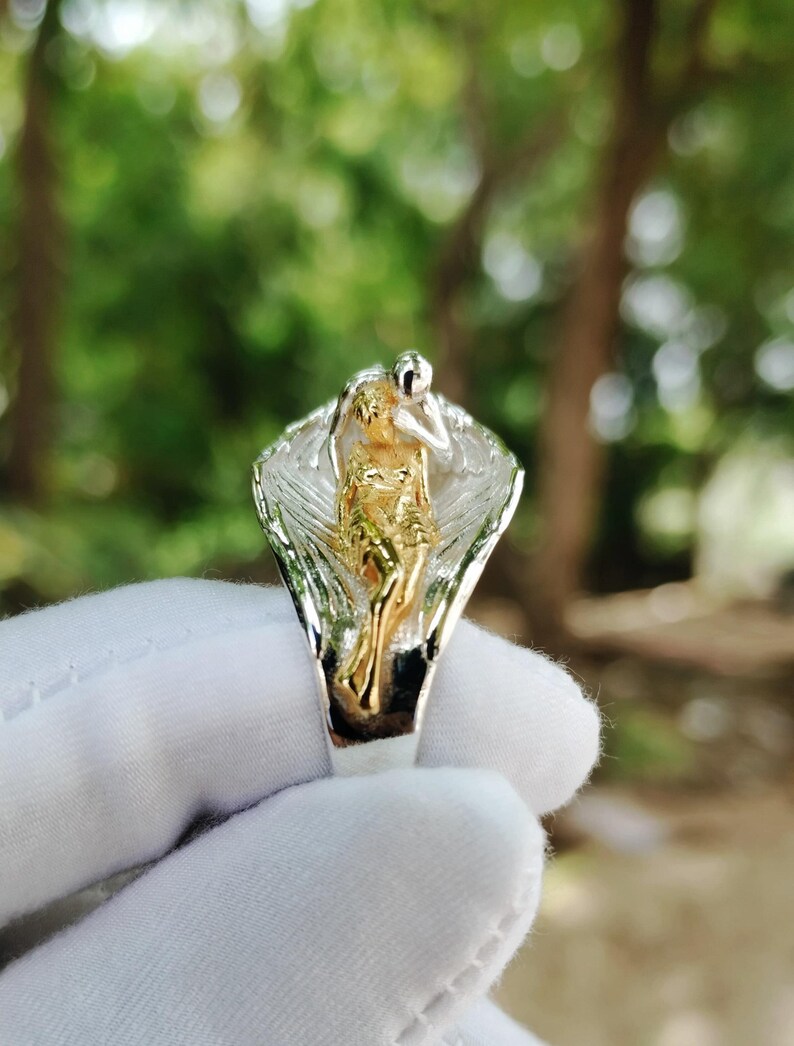 May include: A two-tone ring featuring a gold-colored figure within silver-colored wings. The ring is held by a gloved hand against a blurred green background. The ring's design is intricate and detailed, showcasing a unique artistic style.