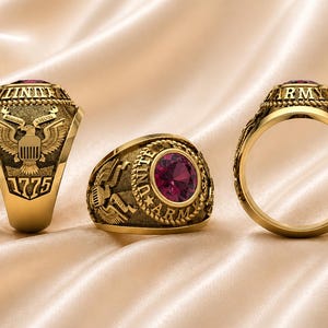 May include: Three gold-colored rings with intricate details. One ring features an eagle emblem and the year 1775. Another ring has a central red gemstone and the word "ARMY". The rings are displayed on a satin surface.