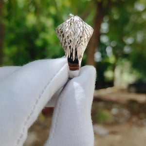 May include: A silver ring with a detailed eagle head design. The ring features a textured surface and a dark interior, held against a blurred green background.