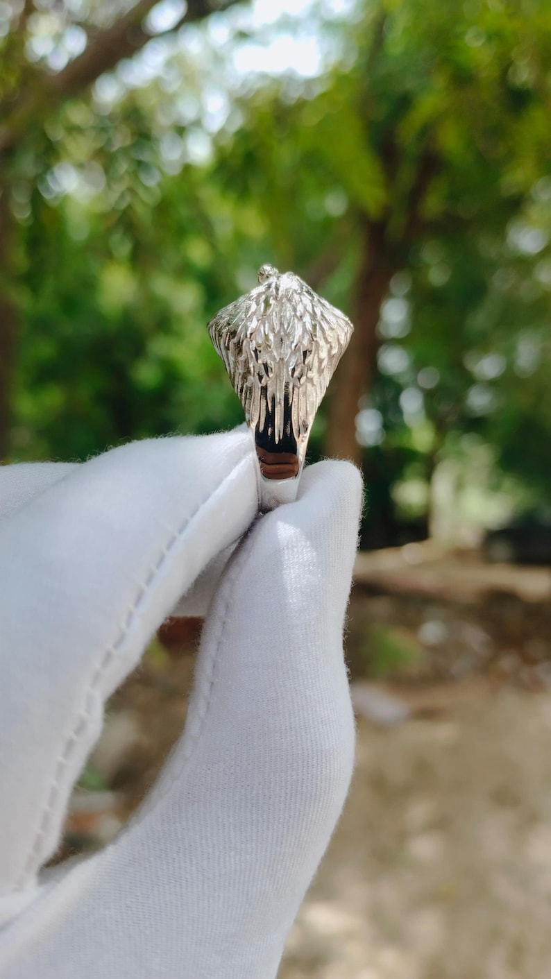 May include: A silver ring with a detailed eagle head design. The ring features a textured surface and a dark interior, held against a blurred green background.