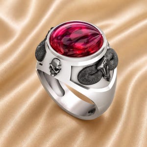 Könnte beinhalten: Silberner Ring mit einem großen, ovalen, roten Edelstein. Der Ring hat ein Totenkopf- und Widderkopf-Design an den Seiten. Der Ring hat eine polierte Oberfläche und ist vor einem goldenen Hintergrund platziert.