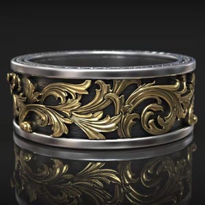 Könnte beinhalten: Ein detaillierter Ring mit einem silbernen Band und einem dunkelgrauen Mittelteil. Die Mitte weist ein verziertes goldenes Blumenmuster auf. Der Ring wird auf einer reflektierenden Oberfläche präsentiert.